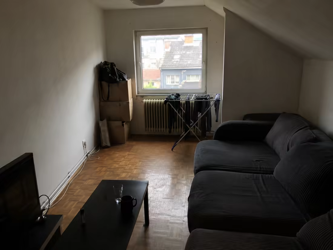 A narrow attic living room with stacked boxes, a drying rack, and a radiator under the window