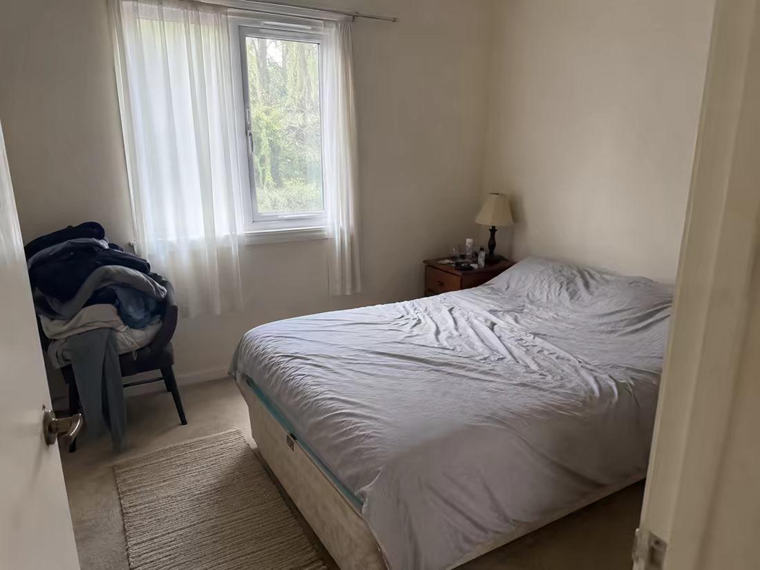 A plain bedroom with rumpled bedding and laundry on a chair