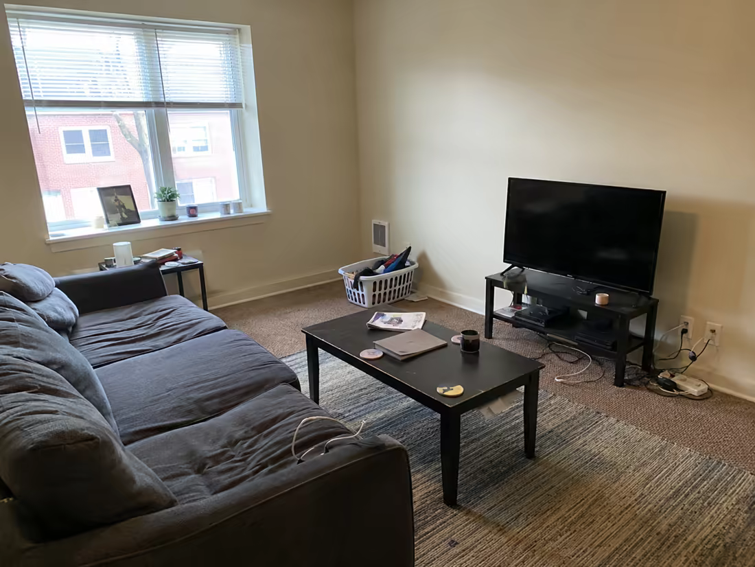 An ordinary living room with a grey sofa, laundry basket, and visible cables, photographed on a phone